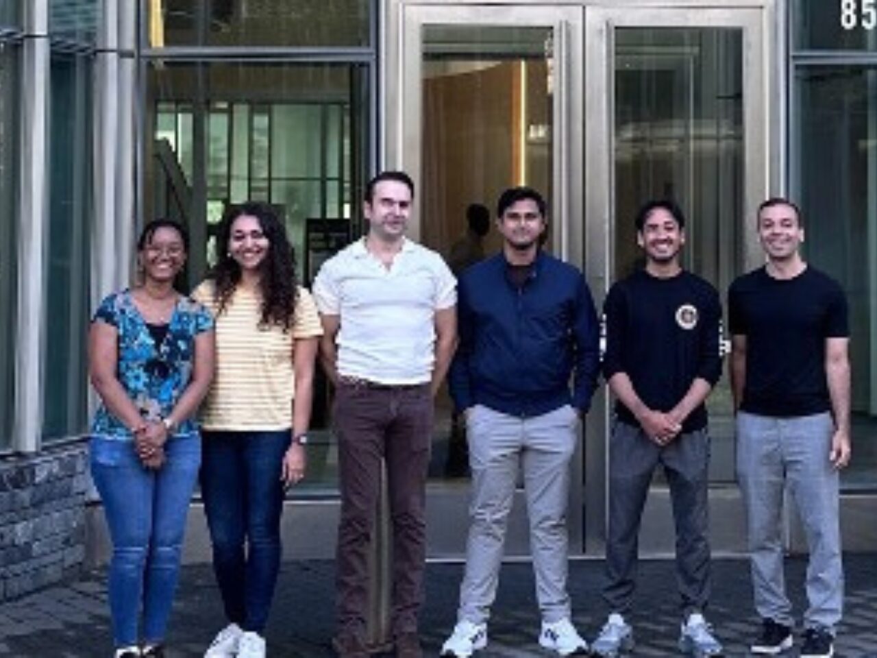 Six people stand in front of a glass building entrance, smiling and posing casually in a group photo.