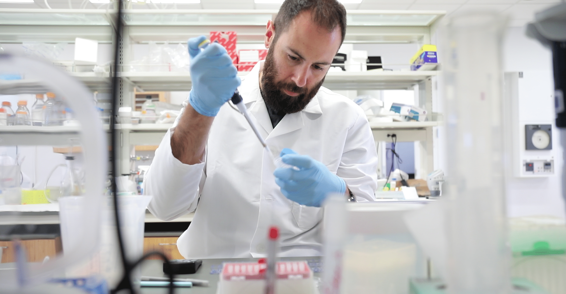 Sami Sauma, a bearded man in a white lab coat, works with research samples in a lab surrounded by shelves of materials