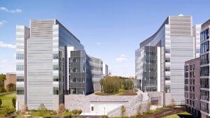 Two modern glass and metal buildings with curved facades, surrounded by greenery and a clear blue sky.