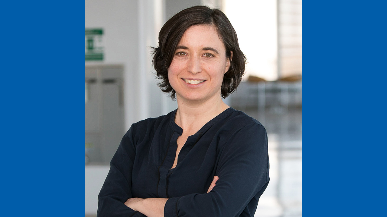 Woman with shoulder-length dark hair smiles confidently, wearing a navy blouse, arms crossed, in a bright, open space.