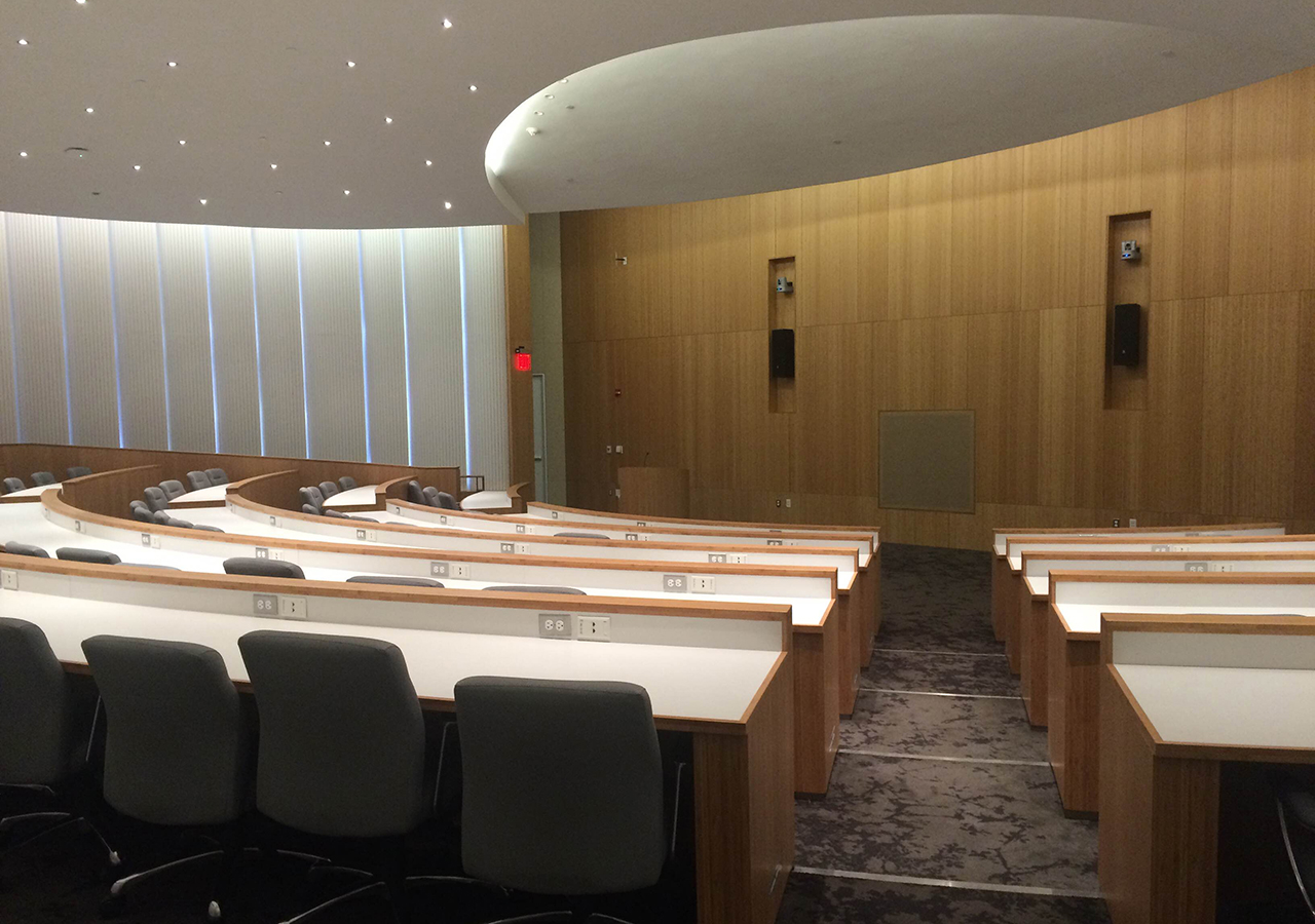 the empty ASRC auditorium is pictured from the rear entrance, looking over curved rows of tabletop seating toward the podium at the front of the room