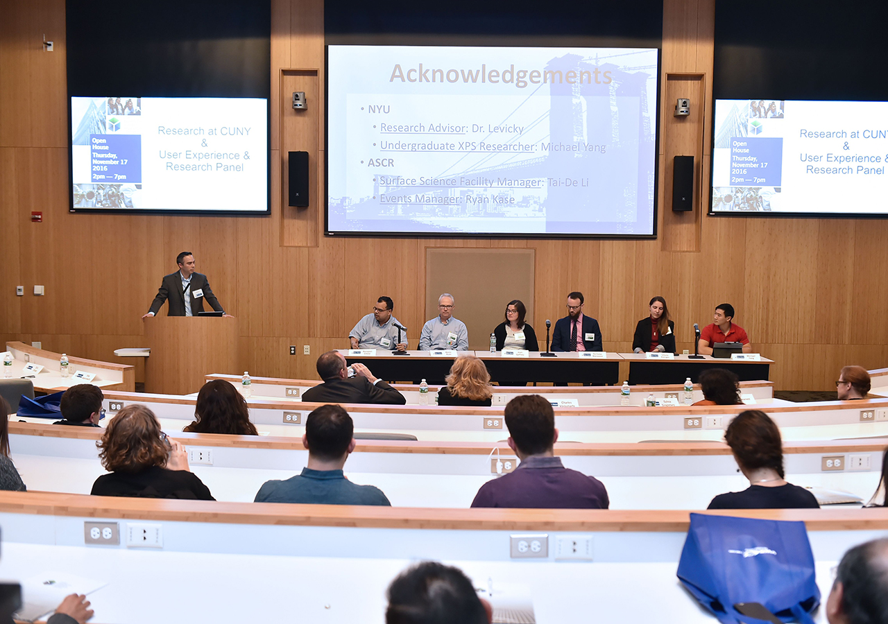 A panel presentation in the auditorium - 6 panelists sit at a long table with the moderator at the podium, seen from the rear center of the room. audience members sit in curved row seating.