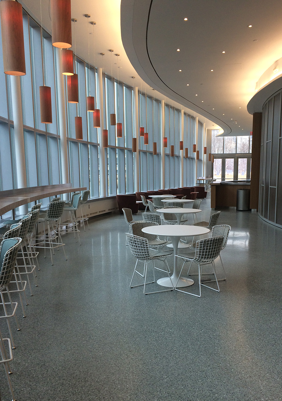 the empty cafe pictured from the main entrance, curving to the right. Pictured are several round white tables with matching chairs and counter seating on the left side of the room. bronze cylindrical pendant lights hang from the ceiling.
