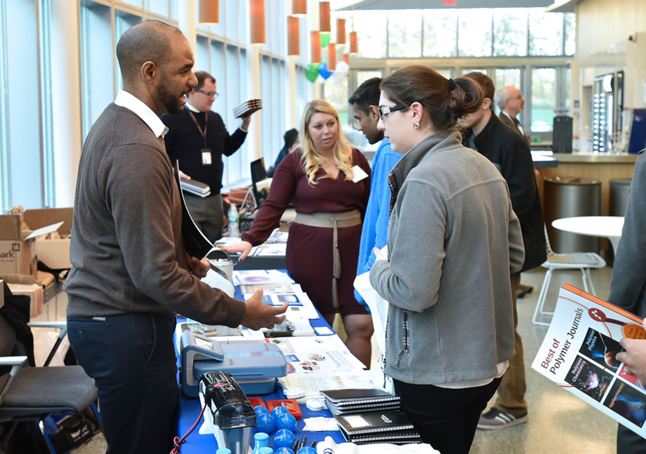 ASRC and GC staff stand at long exhibitor tables and speak to students during an open house event.