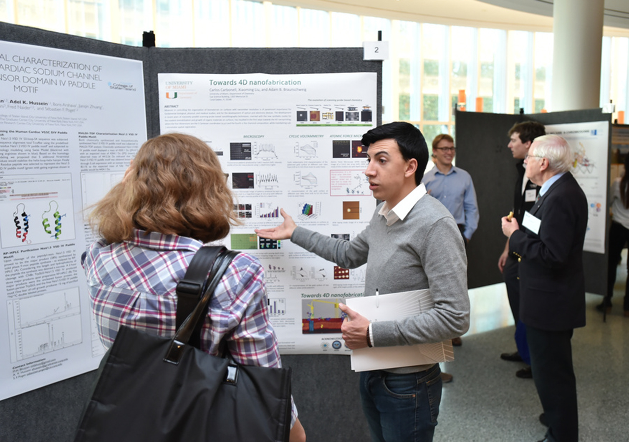 a student researcher gestures to a standing posterboard while discussing their work with an observer during a poster presentation event; other attendees inspect posters and converse in the background