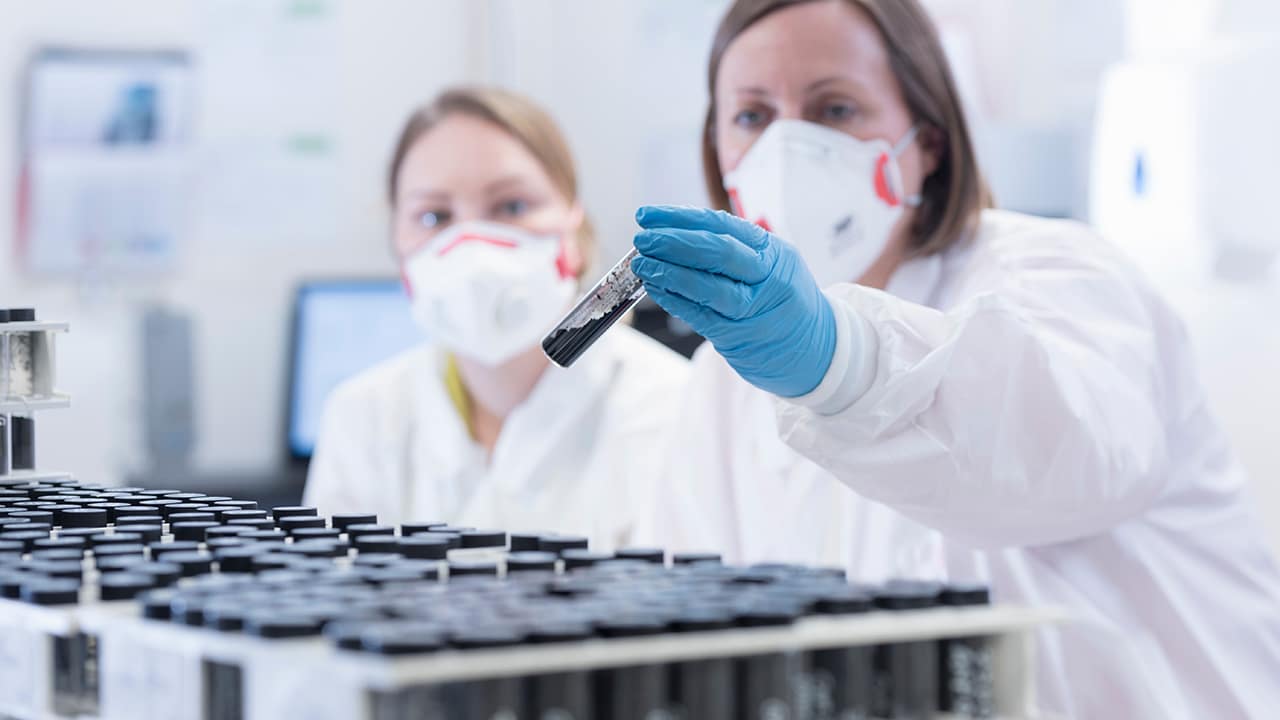 two female researchers in a labr wearing protective gear and looking at a dark material in a test tube