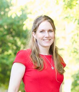 Smiling woman in a red dress stands outdoors with green foliage in the background, wearing a pendant necklace.