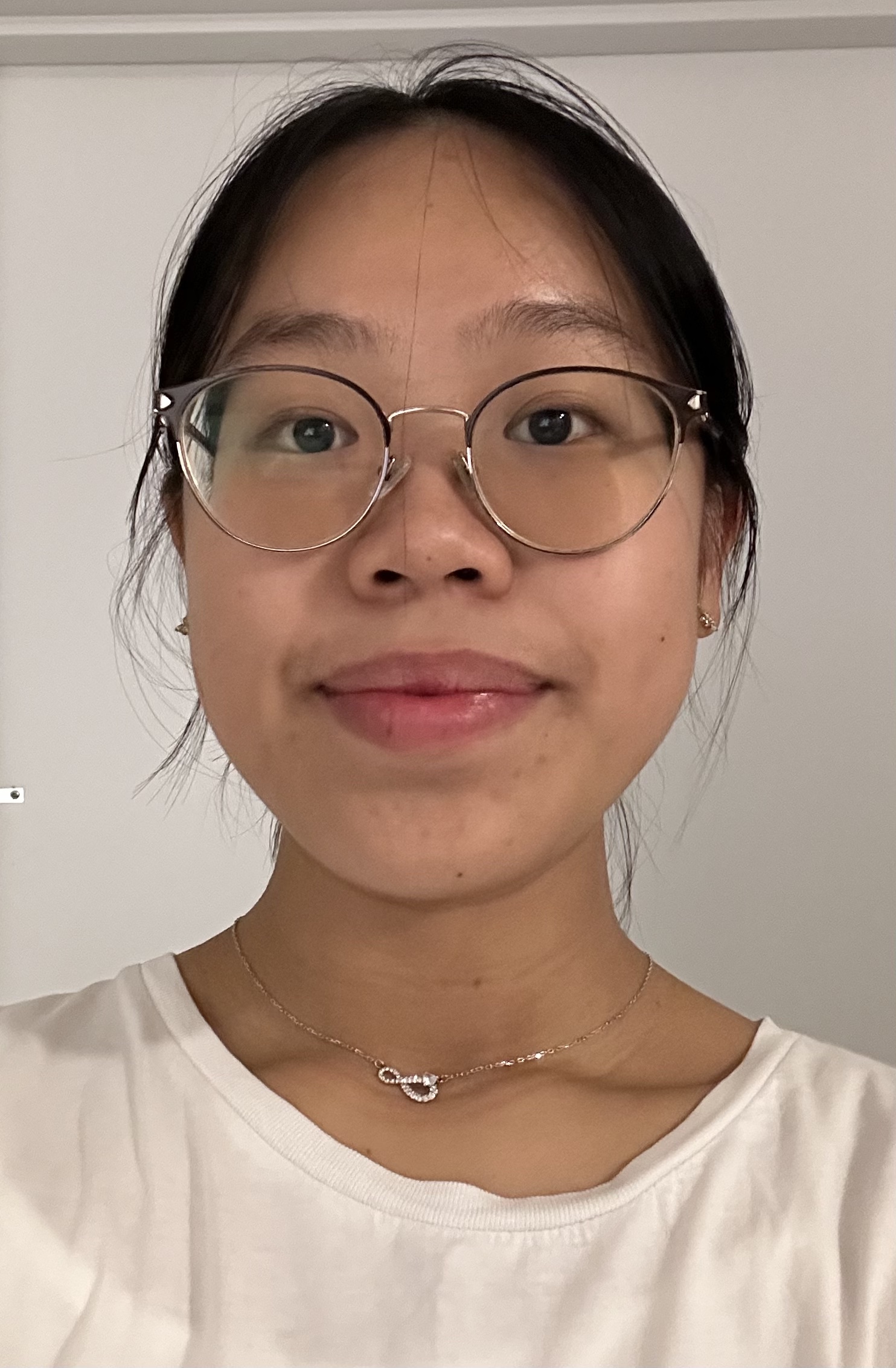 Young woman with glasses and a delicate necklace, smiling against a light background, wearing a white shirt.