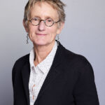 Catherine Cramer wearing glasses, a black blazer over a white polka dot shirt, smiling softly.