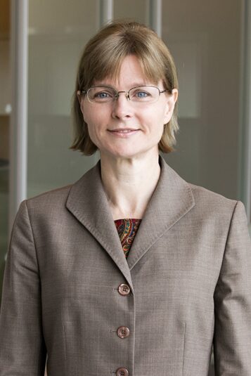A woman with short blonde hair and glasses, wearing a brown suit jacket, stands in an office setting with glass partitions.