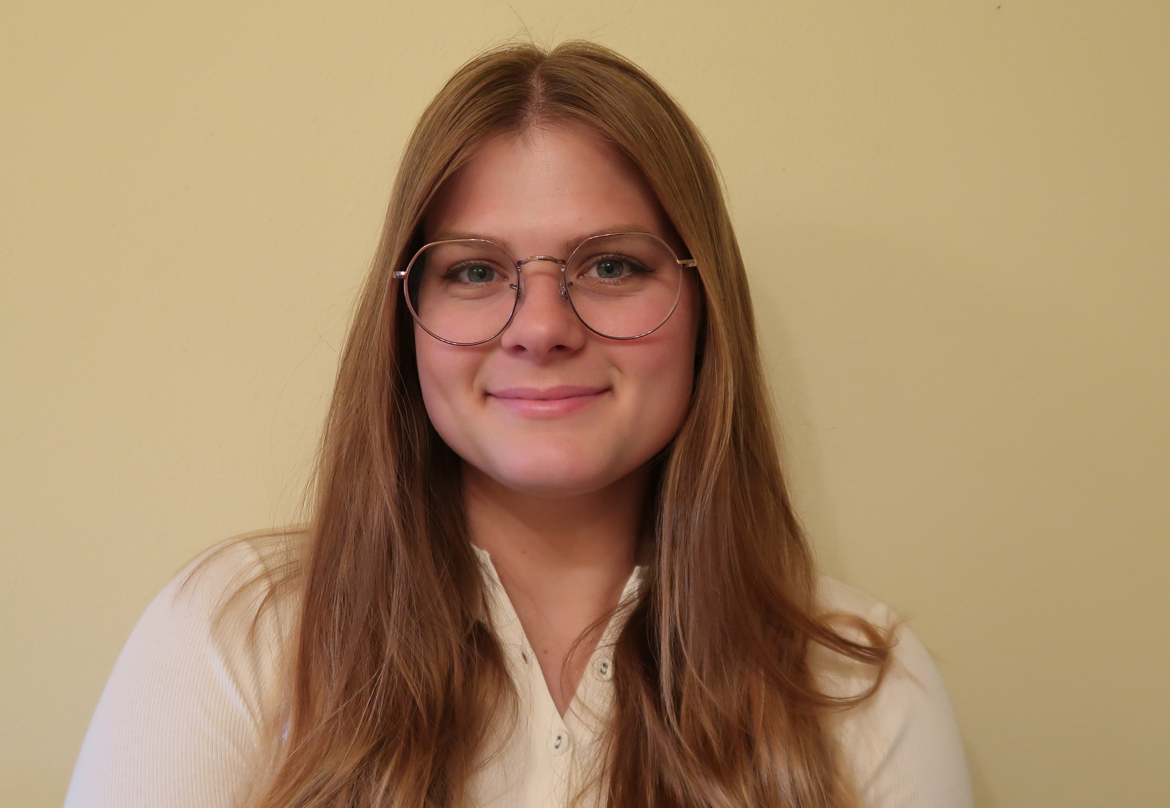 Young woman with long hair and glasses smiles against a yellow background, wearing a white top with buttons.