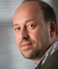 Close-up portrait of a man with a beard and short hair, wearing a sweater, against a soft-focus gray background.
