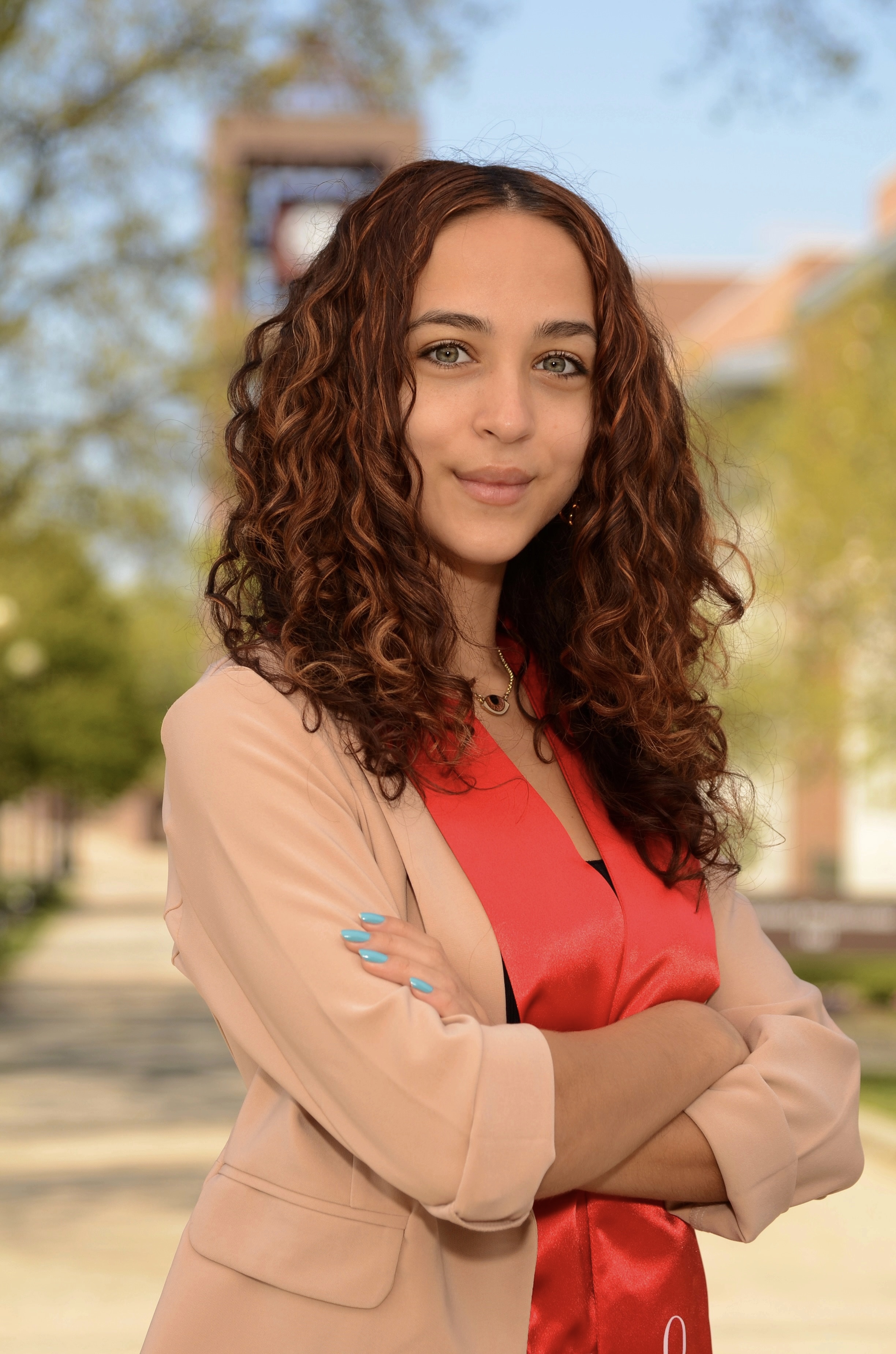 Young woman with curly brown hair poses confidently in a light blazer and red graduation sash, outdoors on a sunny day.