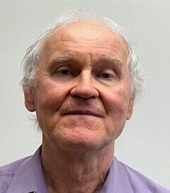 An older man with light gray hair and a slight smile, wearing a purple shirt against a plain background.