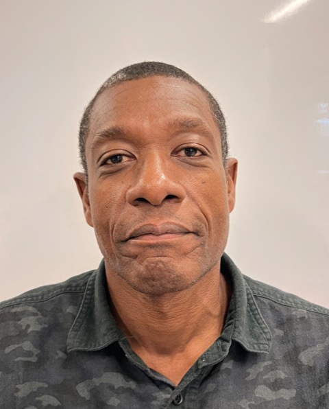 A close-up portrait of a man with short hair, wearing a dark camo-patterned shirt, and a neutral expression.