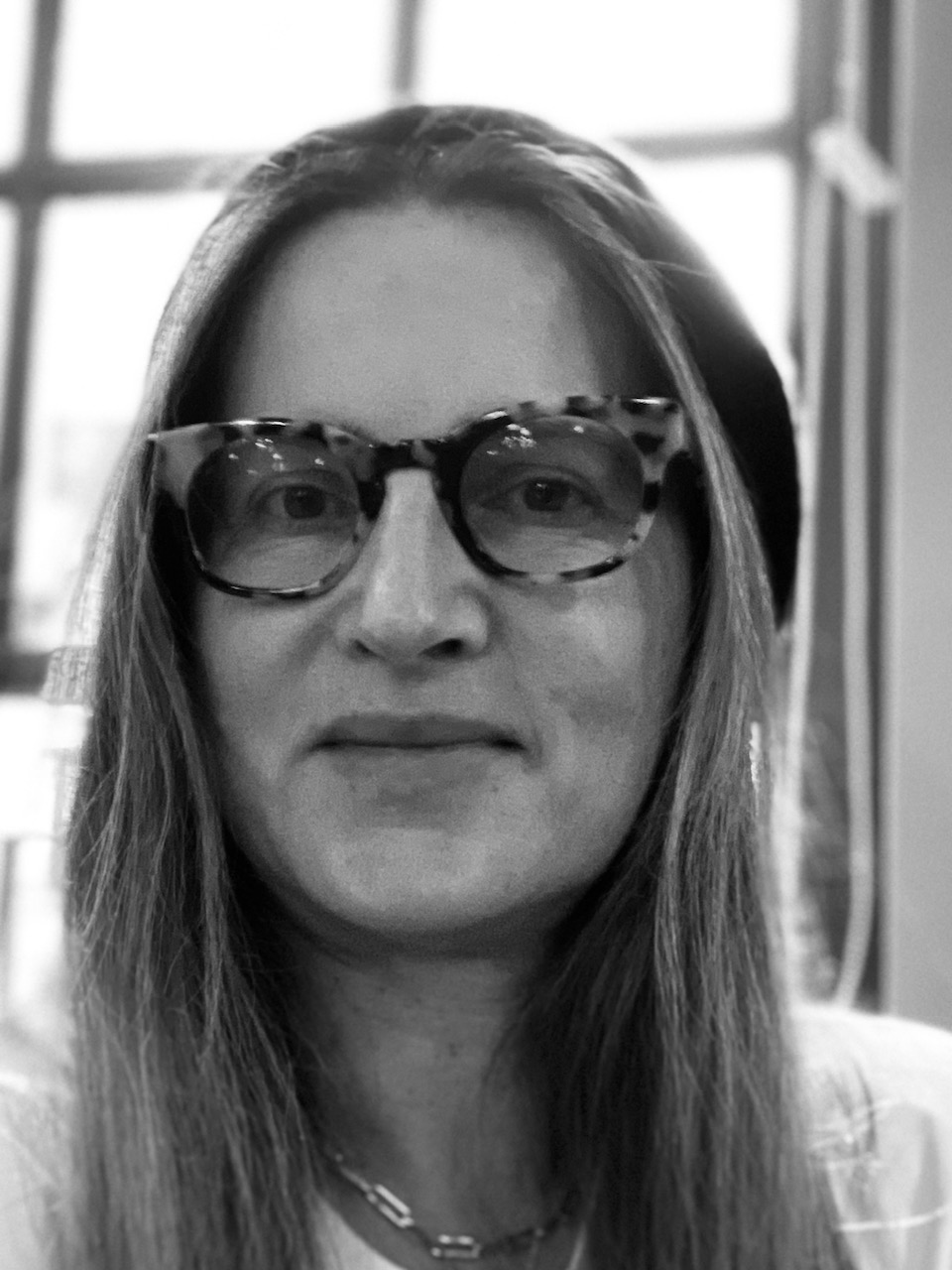 Portrait of a person with long hair wearing glasses and a beanie, smiling softly against a blurred background.