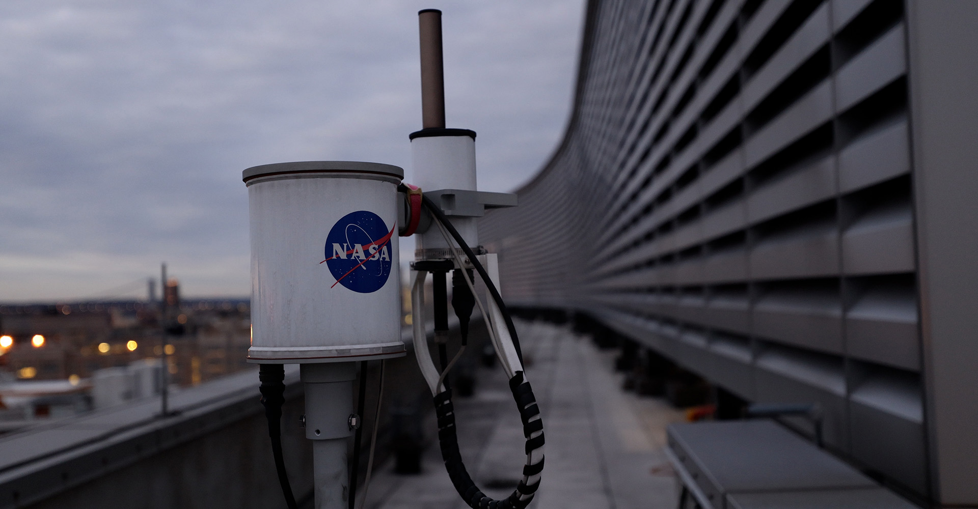 A NASA device with cables on a rooftop, with a blurred cityscape and cloudy sky in the background.