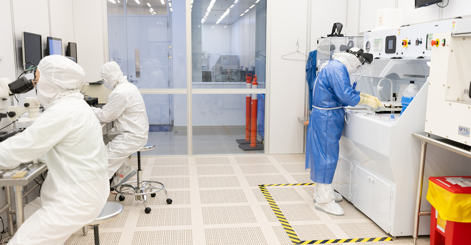 Two scientists in protective suits work in a cleanroom with lab equipment and monitors, focused on research tasks.
