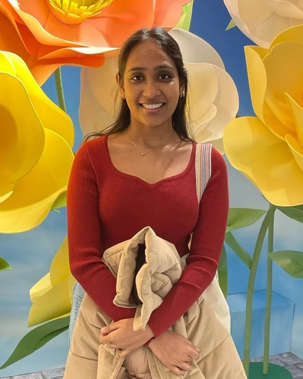 A smiling woman in a red sweater stands against a colorful floral backdrop, holding a light-colored jacket.