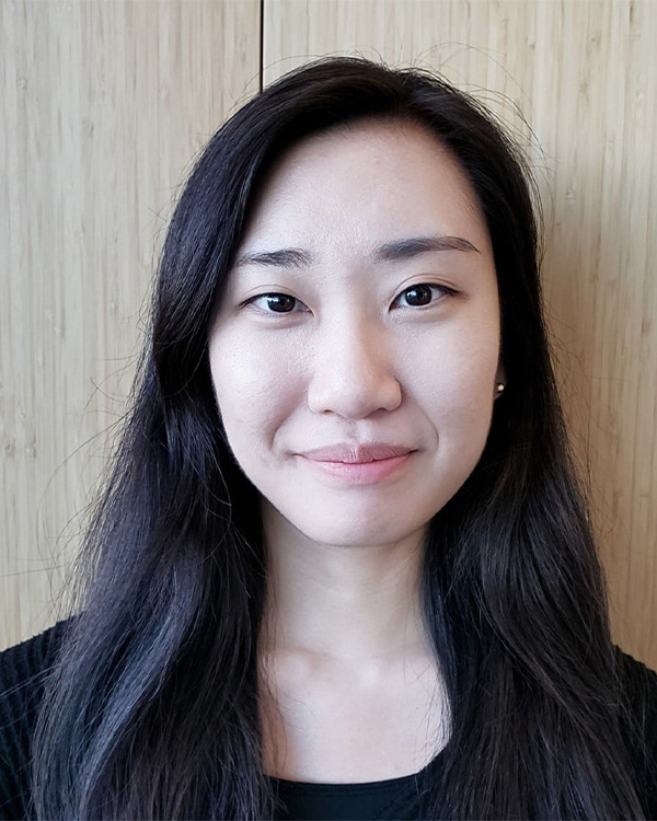 A young woman with long dark hair smiles softly, standing against a light wooden background.