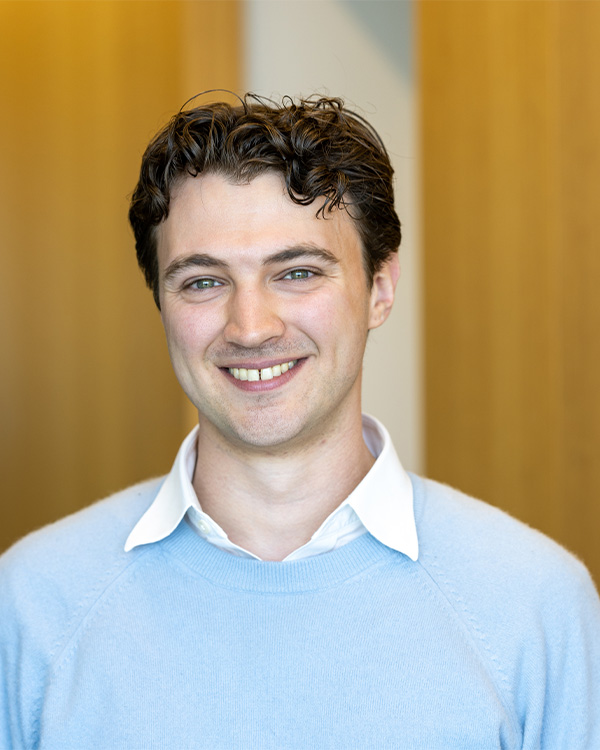 A smiling man with curly hair, wearing a light blue sweater and white collared shirt, stands against a wooden background.