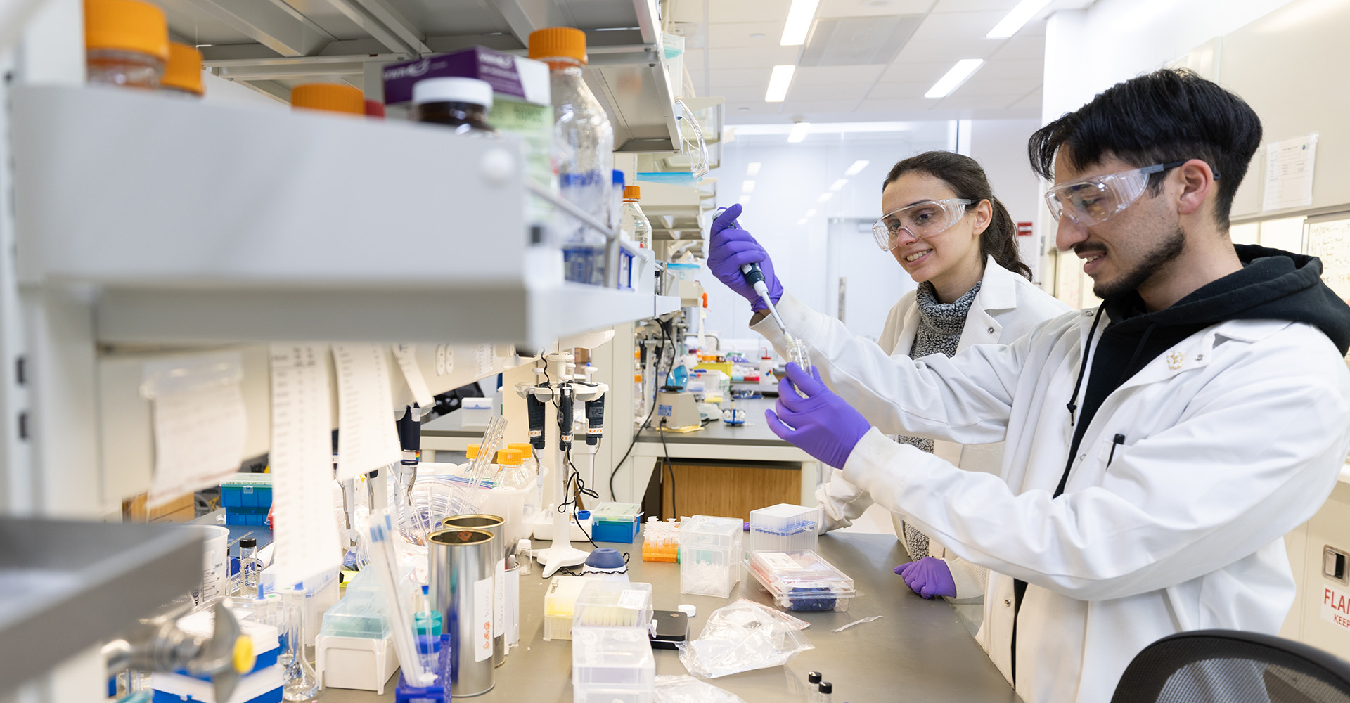 Two lab technicians in white coats and gloves conduct an experiment at a laboratory bench filled with equipment and samples.